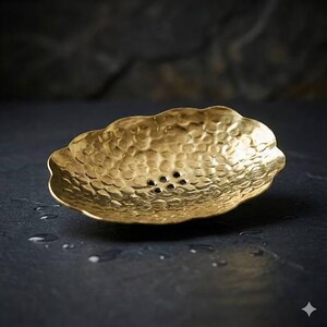 May include: A hammered brass soap dish with a scalloped edge and small drainage holes. The dish has a textured surface and a warm, golden color. The dish is designed to hold a bar of soap.