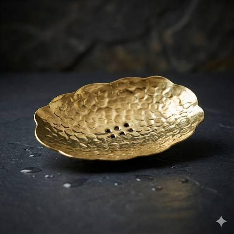 May include: A hammered brass soap dish with a scalloped edge and small drainage holes. The dish has a textured surface and a warm, golden color. The dish is designed to hold a bar of soap.
