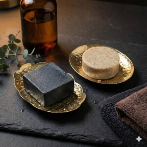 May include: Two gold-colored soap dishes hold a black square soap and a round beige soap. The dishes sit on a dark surface next to two small towels and a brown glass bottle. The soap dishes have a hammered texture.
