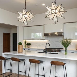 Puede incluir: Dos lámparas colgantes en forma de estrella cuelgan sobre una isla de cocina con encimera de mármol. Las luces tienen un interior blanco y un marco de metal oscuro. La cocina cuenta con gabinetes blancos, taburetes negros y una campana extractora de acero inoxidable.