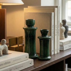 Könnte beinhalten: Zwei dunkelgrüne Kerzenhalter aus Keramik in verschiedenen Größen stehen auf einem dunkelbraunen Regal. Dahinter befinden sich mehrere Bücher mit beigen Einbänden und eine Lampe. Die Kerzenhalter haben eine glänzende Oberfläche.