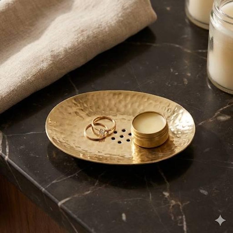 May include: A hammered gold-colored oval tray with two rings and a small round container of balm. The tray has a textured surface and several small holes. The rings are gold with a diamond. The balm container is also gold.