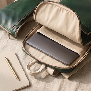 Peut inclure: Un sac à dos en cuir vert foncé avec une doublure intérieure beige, comprenant un compartiment principal contenant un ordinateur portable gris foncé, une pochette zippée, un carnet beige et un stylo doré sur une surface texturée.