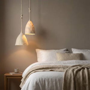 Könnte beinhalten: Zwei cremefarbene, texturierte, glockenförmige Pendelleuchten hängen an dicken, geknoteten Seilen von der Decke. Die Leuchten strahlen ein warmes, sanftes Licht aus und beleuchten den Bereich über einem Bett mit weißer Bettwäsche und einer beigen Strickdecke.