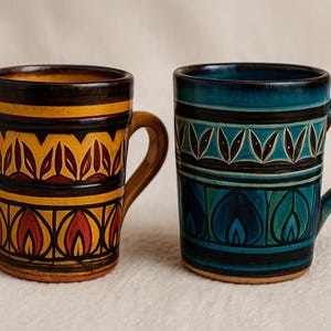 May include: Two ceramic mugs with decorative patterns. One mug is yellow, orange, and black with a leaf design. The other is teal, blue, and black with a leaf and flame design. Both mugs have a curved handle.