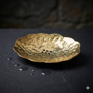 May include: A hammered brass soap dish with a scalloped edge. The dish has a textured surface and several drainage holes. The brass has a warm, golden color. The dish is designed to hold a bar of soap.