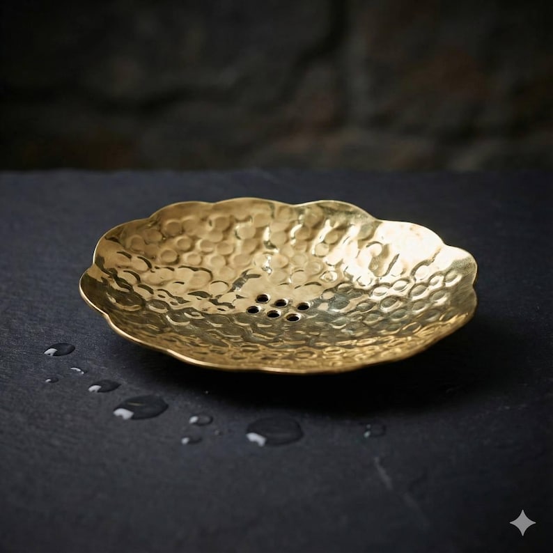 May include: A hammered brass soap dish with a scalloped edge. The dish has a textured surface and several drainage holes. The brass has a warm, golden color. The dish is designed to hold a bar of soap.