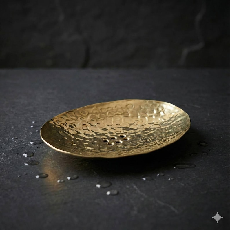 May include: Oval-shaped, hammered brass soap dish. The dish has a textured surface and several drainage holes. The brass has a warm, golden hue, and the dish is set against a dark background.