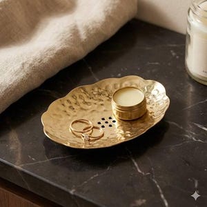 May include: A gold-colored, hammered metal tray with a scalloped edge holds two rings and a small, round tin of balm. The tray sits on a dark, possibly marble, surface. A neutral-colored cloth is partially visible in the background.