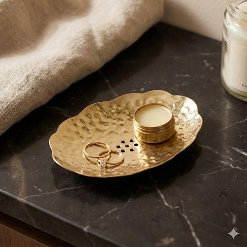 May include: A gold-colored, hammered metal tray with a scalloped edge holds two rings and a small, round tin of balm. The tray sits on a dark, possibly marble, surface. A neutral-colored cloth is partially visible in the background.