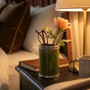Op de afbeelding: Een groene en zilveren pennenhouder met een roos en een bril op een donker houten nachtkastje. Een bruin leren boek en een beige deken liggen ook op het nachtkastje, naast een brandende lamp en een witte mok.