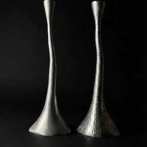 May include: Two silver candlesticks with a modern design stand against a black background. One candlestick has a smooth finish, while the other has a textured, hammered appearance. The bases are flared, and the stems curve gracefully.