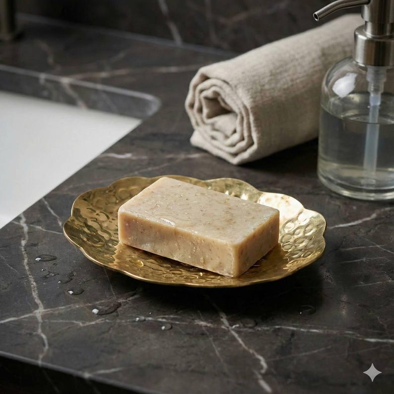 May include: A gold-colored, scalloped soap dish holds a rectangular bar of soap. A rolled-up beige towel and a clear glass soap dispenser with a silver pump are in the background. The soap dish sits on a dark, marble-patterned surface.