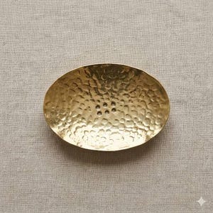 May include: Oval-shaped, hammered brass soap dish. The dish has a textured surface and six drainage holes. The brass has a warm, golden hue. The soap dish is designed to hold a bar of soap.