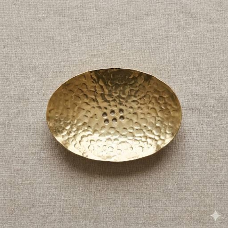 May include: Oval-shaped, hammered brass soap dish. The dish has a textured surface and six drainage holes. The brass has a warm, golden hue. The soap dish is designed to hold a bar of soap.