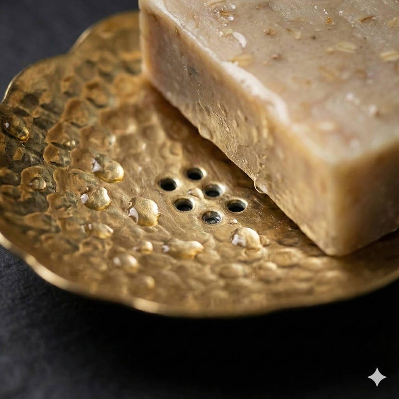 May include: A gold-colored, hammered metal soap dish with a floral design, holding a bar of beige soap. The soap dish has small drainage holes and is covered in water droplets. The soap has visible grains and a slightly uneven surface.