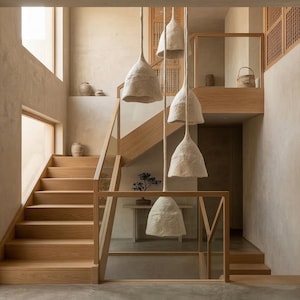 May include: A multi-tiered pendant light fixture featuring five off-white, textured, bell-shaped shades suspended at varying heights. The light fixture hangs in a bright, open-concept interior space with light wood stairs and beige walls.
