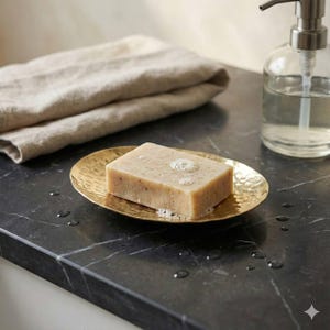 May include: A rectangular bar of soap with bubbles sits on a hammered gold-colored soap dish. A clear glass soap dispenser with a silver pump and a folded beige towel are in the background. The soap dish is on a dark countertop.