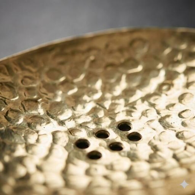 May include: Close-up of a hammered brass soap dish. The dish has a textured surface with a series of small, evenly spaced drainage holes. The brass has a warm, golden hue, reflecting light.