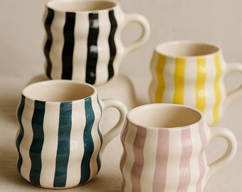 May include: Four ceramic mugs with wavy vertical stripes in black, yellow, teal, and pink. Each mug has a white background and a rounded handle. The mugs are a unique and stylish addition to any kitchen.