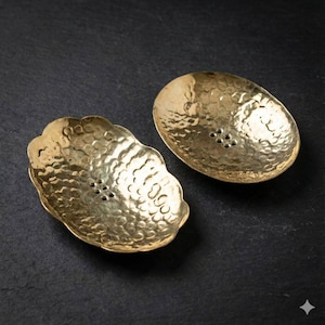 May include: Two hammered brass soap dishes. One dish has a scalloped edge, the other is oval. Both have a textured surface and drainage holes. The dishes are displayed on a dark surface.