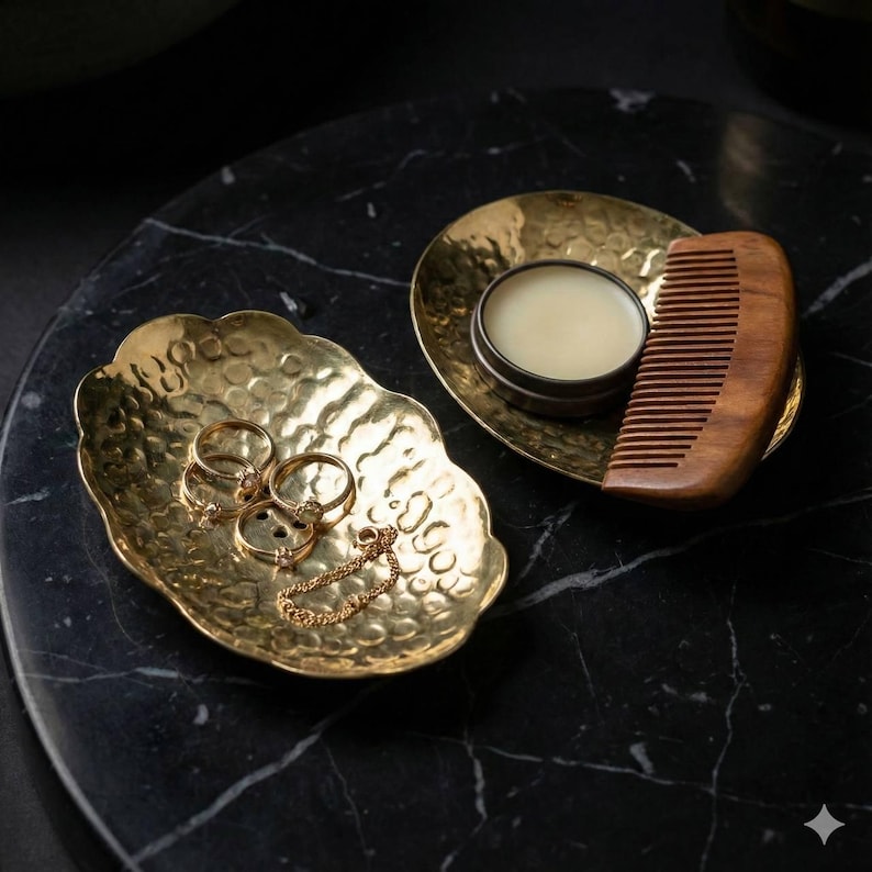 May include: Two hammered gold-colored metal trays on a black marble surface. One tray holds rings and a bracelet. The other holds a small tin of balm and a wooden comb. The trays have a textured, dimpled surface.