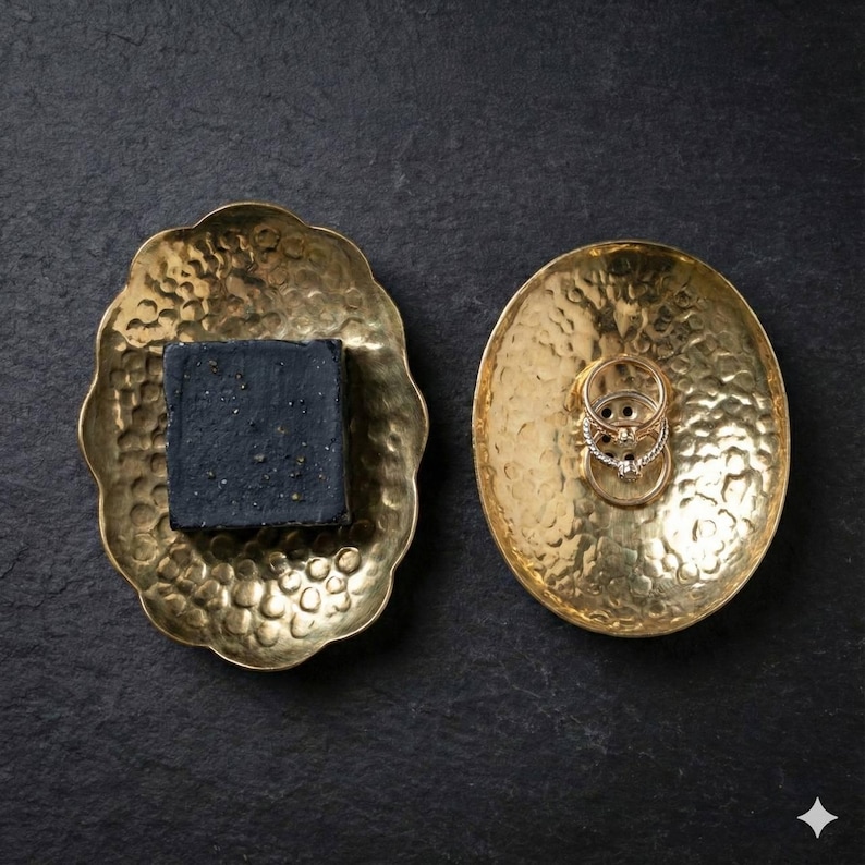 May include: Two hammered brass trays on a dark surface. One tray holds a dark gray bar of soap, the other holds several rings. The trays have a textured surface and a decorative, organic shape. The rings are gold and silver.