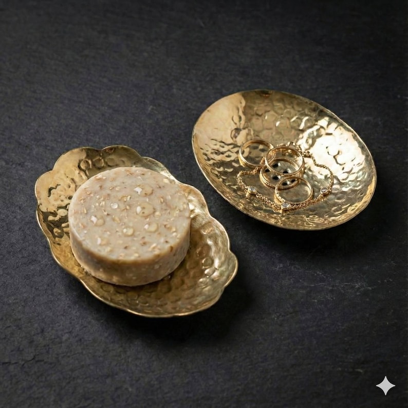 May include: Two gold-colored, hammered metal trays on a dark surface. One tray holds a round bar of soap, the other holds gold rings and a bracelet. The trays have a decorative, organic shape and a textured surface.