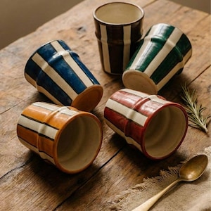 May include: A collection of ceramic cups with vertical stripes in various colors, including blue, green, orange, red, and brown. The cups are arranged on a wooden surface with a spoon and a sprig of rosemary.