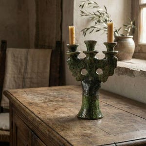 May include: A green ceramic candelabra with three candle holders, each holding a lit, yellow candle. The candelabra is on a wooden table. The background includes a window, a chair, and a vase with greenery.