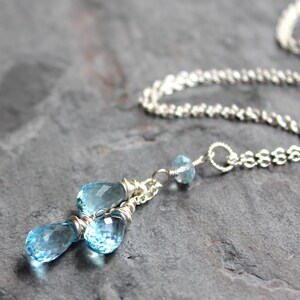 May include: A delicate silver necklace featuring three light blue teardrop-shaped gemstones. The gemstones are wire-wrapped and suspended from a delicate silver chain. A smaller blue gemstone is also present. The necklace is displayed on a dark gray surface.