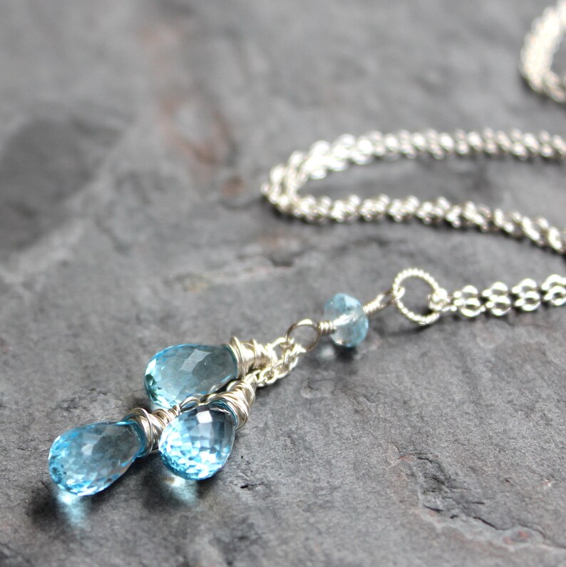 May include: A delicate silver necklace featuring three light blue teardrop-shaped gemstones. The gemstones are wire-wrapped and suspended from a delicate silver chain. A smaller blue gemstone is also present. The necklace is displayed on a dark gray surface.