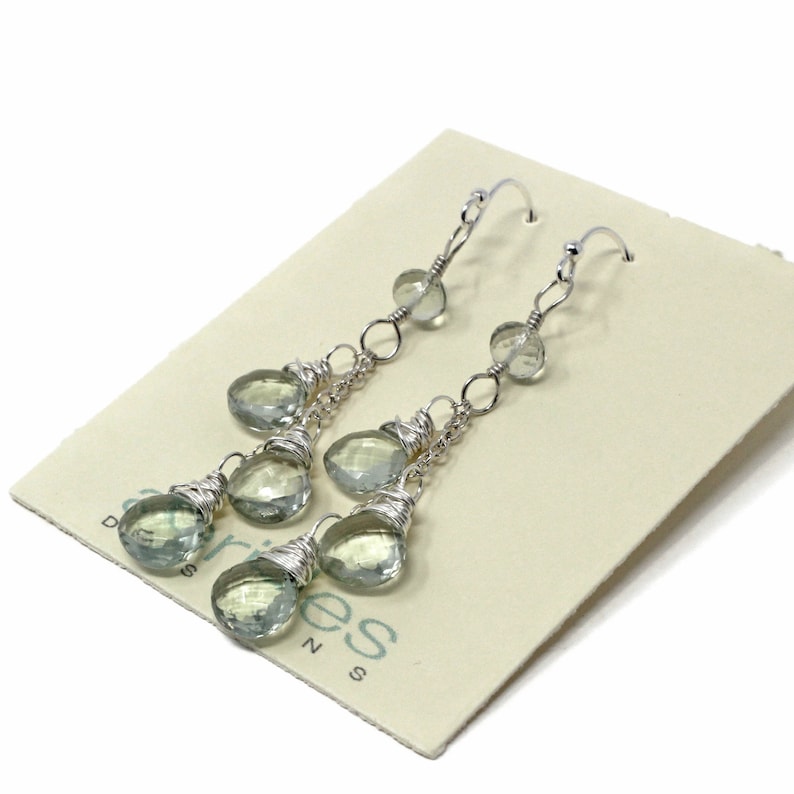 May include: Silver dangle earrings with multiple light green gemstone drops. The earrings are on a white card with the text "Aries Designs" printed on it.