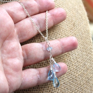 May include: A delicate silver necklace featuring a cascade of blue gemstone pendants. The necklace has a fine chain and three teardrop-shaped blue gemstones, with a smaller blue stone above them. The gemstones are wire-wrapped.