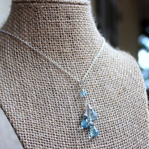 May include: A delicate silver necklace featuring a cascade of blue teardrop-shaped gemstones. The necklace is displayed on a burlap-textured surface, showcasing the sparkling blue stones and the fine silver chain.