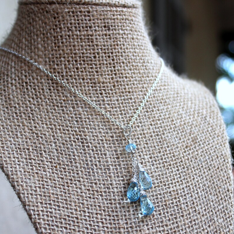 May include: A delicate silver necklace featuring a cascade of blue teardrop-shaped gemstones. The necklace is displayed on a burlap-textured surface, showcasing the sparkling blue stones and the fine silver chain.