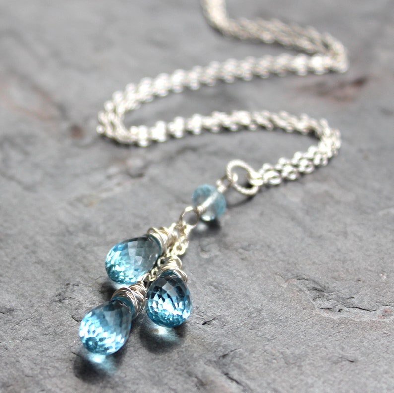 May include: A silver necklace with three blue teardrop-shaped gemstones. The gemstones are wire-wrapped and dangle from a delicate chain. The necklace has a twisted silver chain. The background is a dark gray surface.