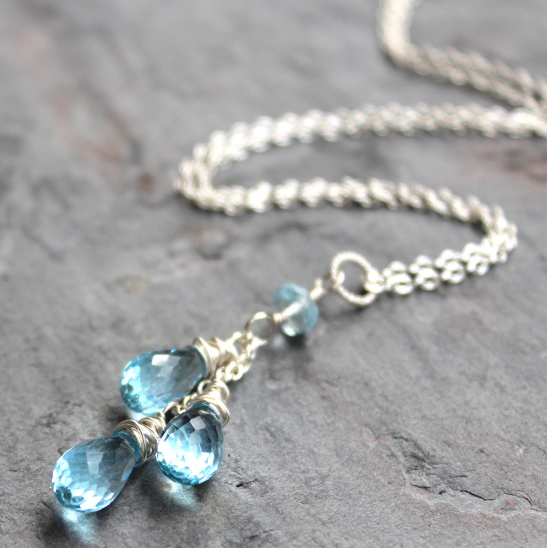 May include: A silver necklace featuring a delicate chain and three blue teardrop-shaped gemstones. The gemstones are faceted, catching the light, and are wire-wrapped. The necklace is displayed on a dark gray surface.