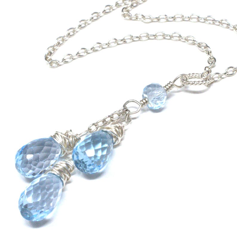 May include: A silver necklace featuring a delicate chain and three teardrop-shaped, faceted, light blue gemstones. The gemstones are wire-wrapped and suspended from the chain, creating a layered effect. A smaller, round gemstone is also present.