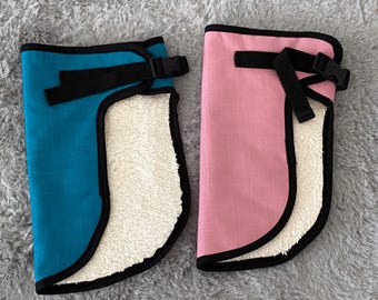 May include: Two protective leg wraps, one teal and one pink, are displayed. Each wrap has a black trim, a black strap with a buckle, and a white, fluffy interior. The wraps are designed to protect the legs.