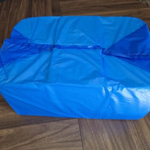 May include: A rectangular, bright blue plastic bag, folded and sealed. The bag is a solid, vibrant blue with darker blue accents where the folds and seams are. The bag is sitting on a wooden surface.