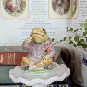 May include: A ceramic figurine of a frog dressed in a mauve jacket and tan breeches, sitting cross-legged on a green lily pad base. The frog has a light green face, dark eyes, and holds a small orange object in one hand while resting the other.