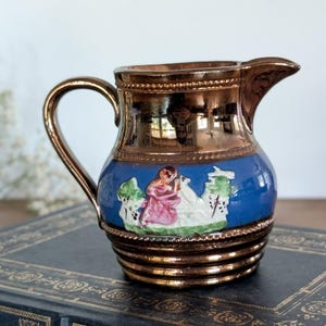 May include: A small ceramic creamer featuring a metallic copper finish on the top and bottom sections. The center band is a vibrant blue with a raised, hand-painted relief depicting a figure in a pink garment kneeling beside a white animal.