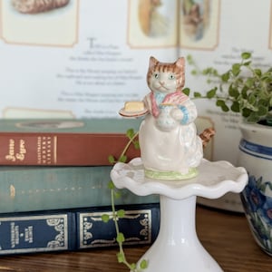 May include: A ceramic cat figurine dressed in a pink scarf and white apron, holding a small plate with a pat of butter and a blue and white patterned pitcher. The cat has brown tabby markings on its head and tail and stands on a white pedestal.