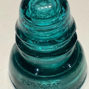 May include: A vintage teal glass electrical insulator with a stepped, dome-shaped profile. The base features the embossed text HEMINGRAY. The glass is translucent with a smooth, glossy finish and a tiered design narrowing toward the top.