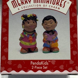 May include: A Merry Miniatures PendaKids 2-Piece Set box featuring two small figurines. One figurine wears a pink dress with a colorful necklace, and the other wears a blue and orange outfit while holding a small doll. The box has a red border with hearts.