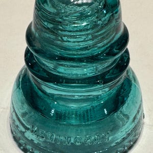 May include: A teal-colored glass electrical insulator with a rounded top and tiered, ridged sides. The base features the embossed text HEMINGRAY. The object has a smooth, translucent finish and a wide, circular bottom edge with a decorative beaded pattern.