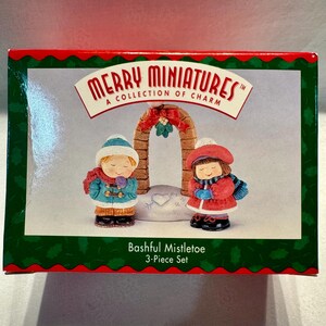 May include: A Merry Miniatures 3-piece set box featuring a Bashful Mistletoe collection. The box displays two small figurines of children in winter clothing standing before a brick archway with a red bow and mistletoe, all on a snowy base.