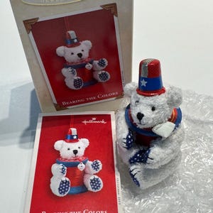 May include: A white plush teddy bear ornament wearing a red, white, and blue top hat with a star. The bear sits on a red spool of thread with blue ends. The bear has blue and white star-patterned paw pads. Includes a Hallmark box and card.
