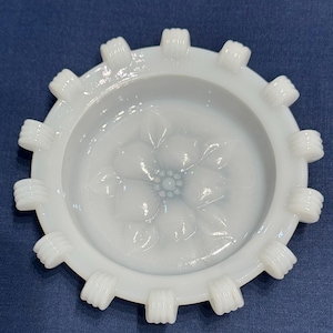 Può includere: Un posacenere bianco e circolare con un motivo floreale al centro. Il posacenere ha una serie di sporgenze rettangolari attorno al bordo esterno. Il posacenere è realizzato in vetro o ceramica opaca.